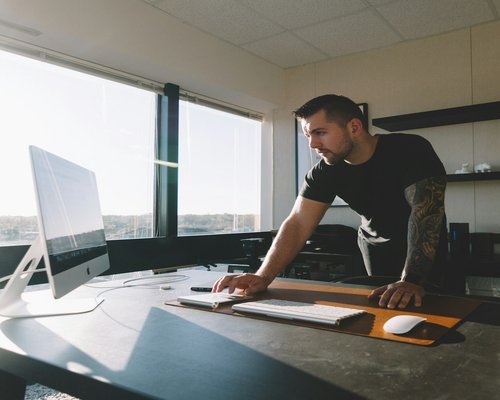 A side view of a young professional stand by an office table near large window panels emphasizing workplace ergonomics